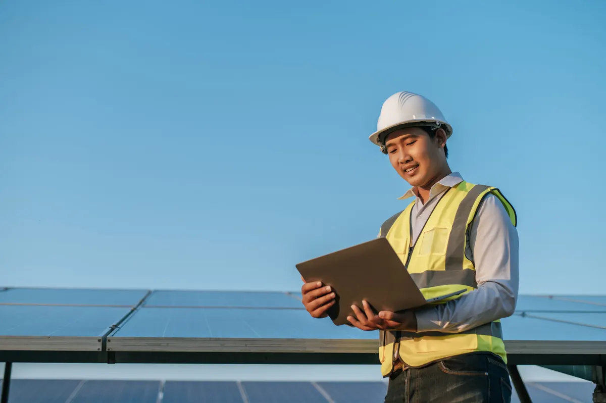 A supervisor in a white helmet checking a solar panel station with a laptop